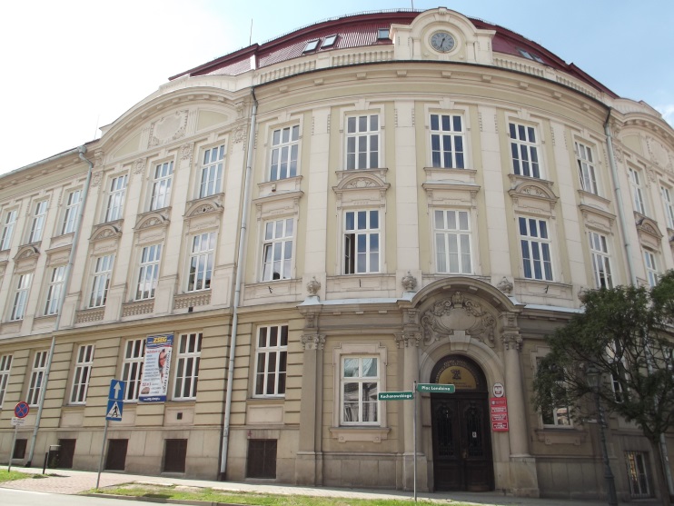 Trzykondygnacyjny budynek o zaokrąglonej fasadzie z licznymi prostokątnymi oknami. Wejście główne z ozdobnym portalem. Nad drzwiami godło państwowe. Przy budynku znaki drogowe i tablice informacyjne.
