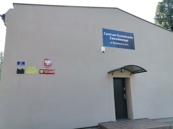 Jednokondygnacyjny budynek o jasnej elewacji. Na ścianie widoczne trzy tablice informacyjne oraz szyld z napisem 'Centrum Kształcenia Zawodowego w Bażanowicach'. Wejście z daszkiem nad drzwiami.