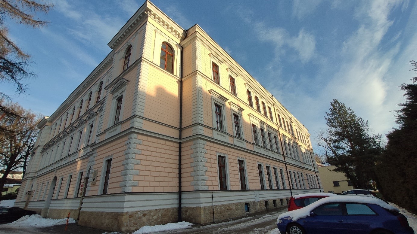 Budynek murowany, trzykondygnacyjny, o jasnej elewacji z regularnie rozmieszczonymi oknami. Widok narożny. Na pierwszym planie zaparkowane samochody. Na podłożu śnieg. W tle drzewa i niebo