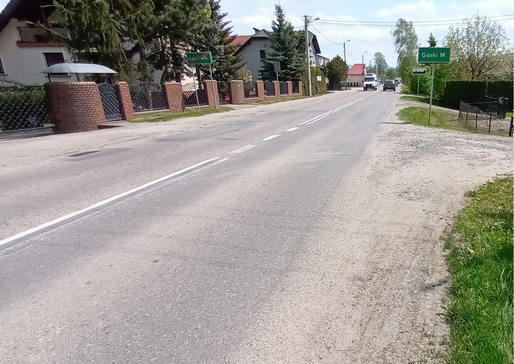 Droga asfaltowa w terenie zabudowanym, po obu stronach chodniki i domy jednorodzinne z ogrodzeniami. Po prawej stronie znak z nazwą miejscowości „Górki M.”