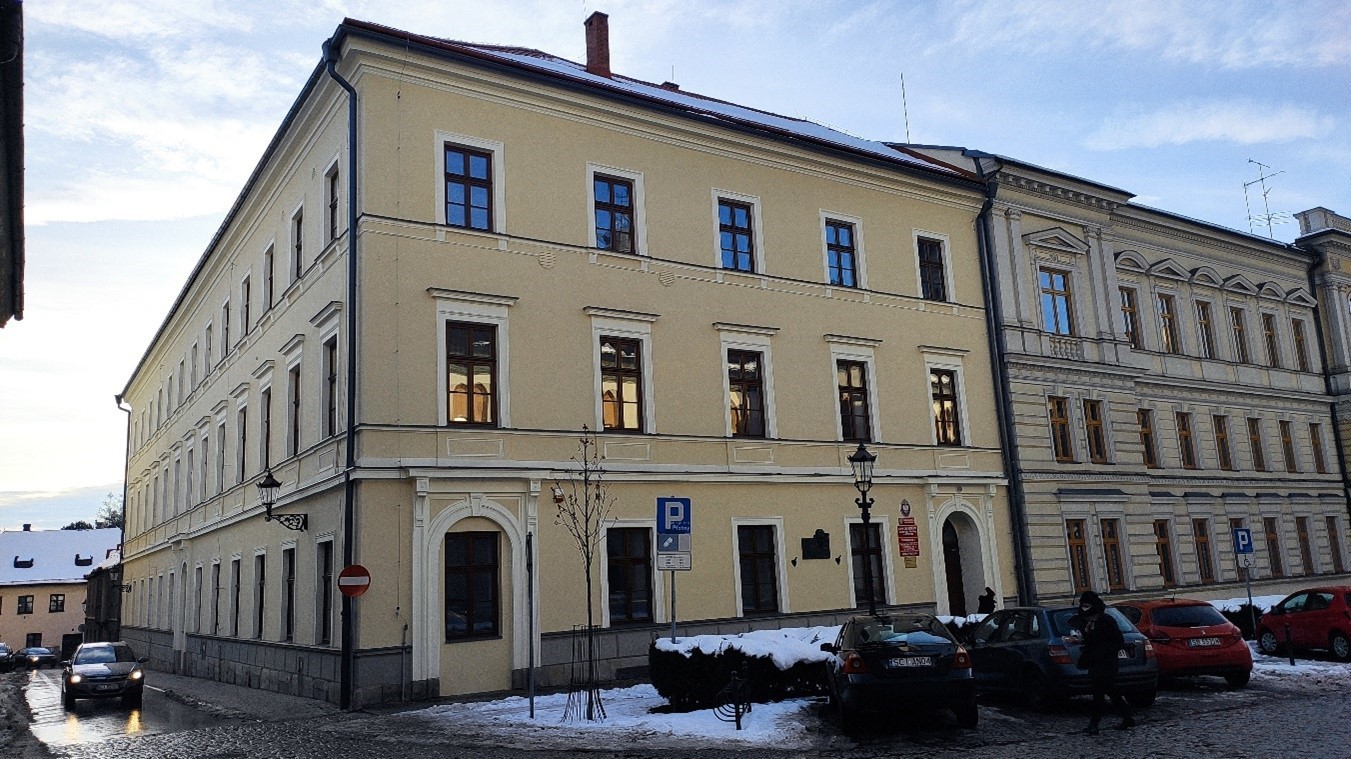 Dwukondygnacyjny budynek o jasnej elewacji z prostokątnymi oknami. Widok narożny. Na parterze tablice informacyjne. Przed budynkiem zaparkowane samochody. Na chodniku zalega śnieg.