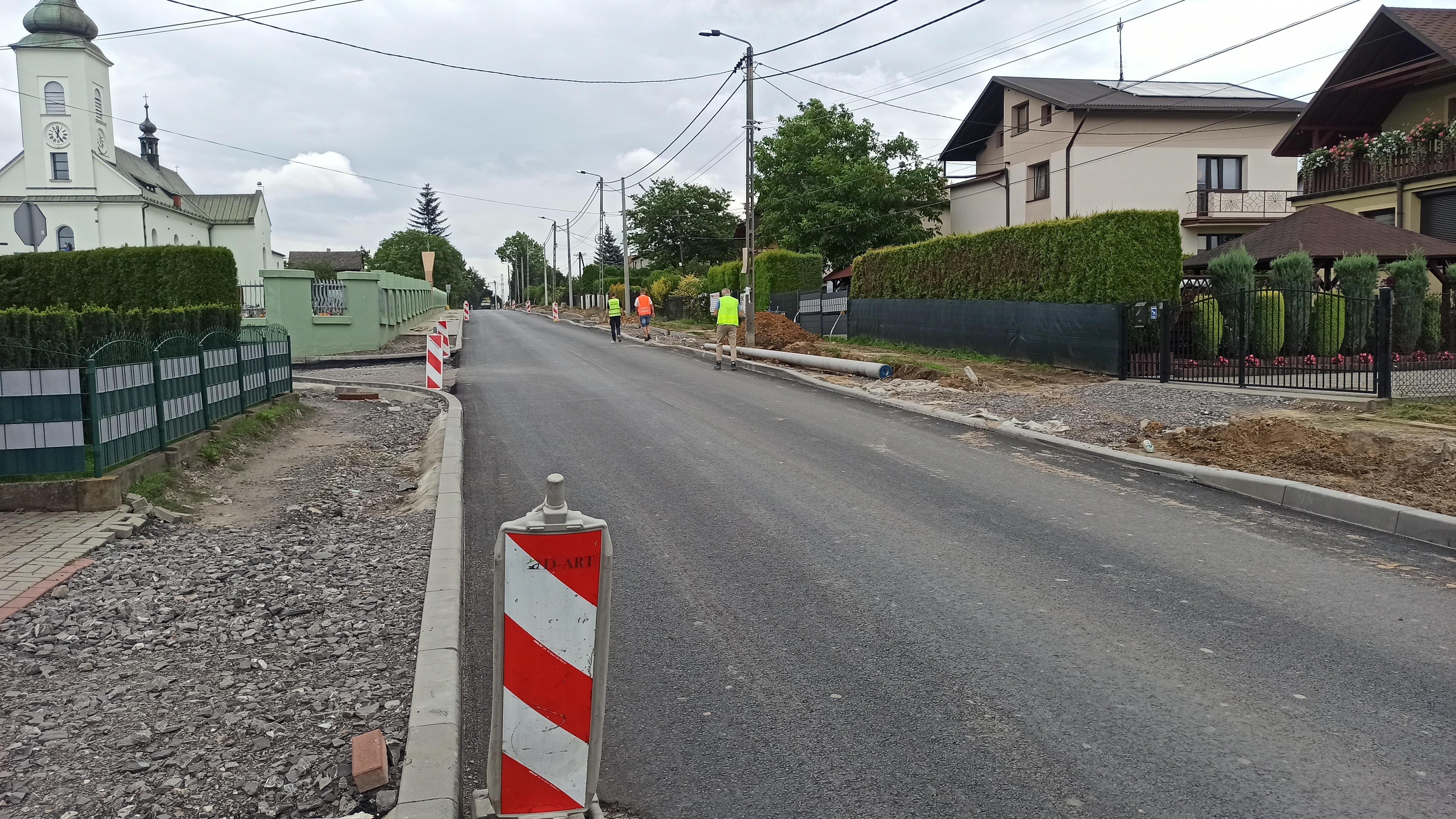 Widok na przebudowywaną drogę w terenie zabudowanym. Na jezdni położona jest nowa nawierzchnia asfaltowa, a po obu stronach trwają prace przy chodnikach i poboczach. Wzdłuż drogi ustawiono oznakowanie rob&oacute;t, a w tle widoczni są pracownicy oraz zabudowania, w tym kości&oacute;ł.