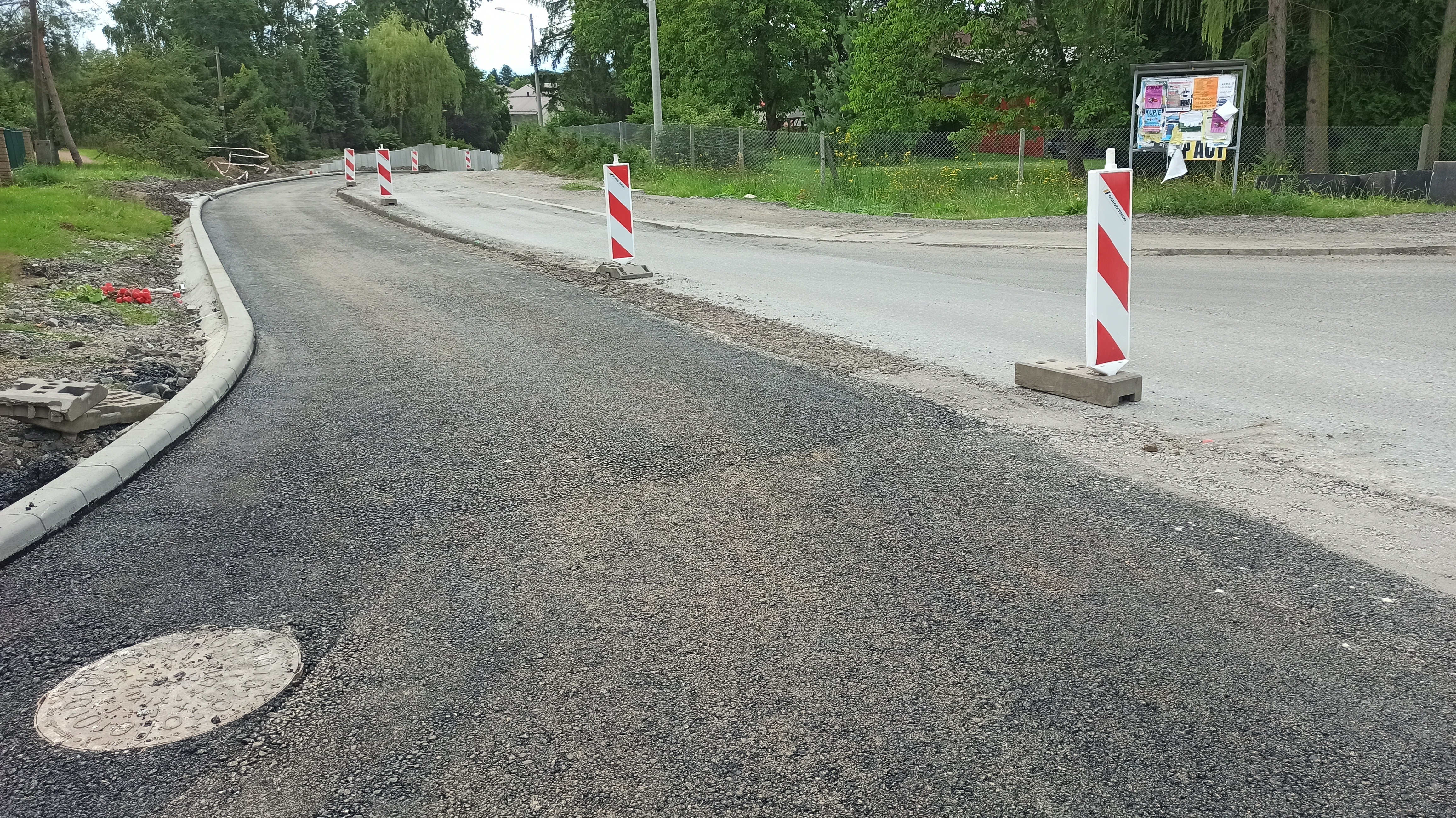 Widok na remontowany odcinek drogi. Na części jezdni położona jest nowa nawierzchnia asfaltowa, a obok trwają dalsze prace drogowe. Wzdłuż trasy ustawiono biało-czerwone tablice ostrzegawcze, a po lewej stronie widoczny jest nowy krawężnik i przygotowane pobocze.