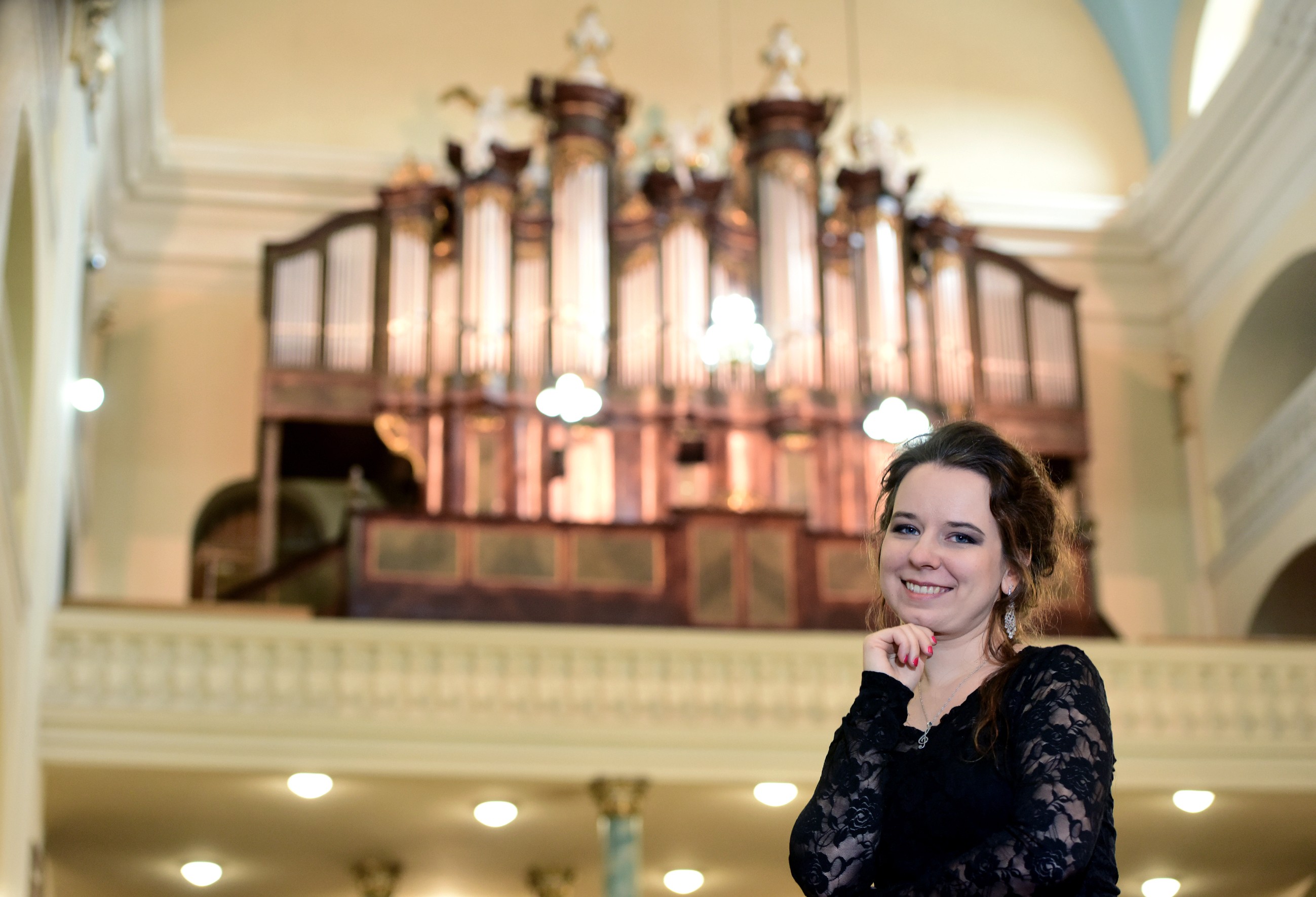 Wnętrze kościoła z dużym prospektem organowym w tle. Na pierwszym planie Ewelina Bachul-Cienciała w czarnej, koronkowej sukni, uśmiechnięta, z ręką opartą o brodę. W tle widoczne kolumny, balustrada i oświetlenie sufitowe.