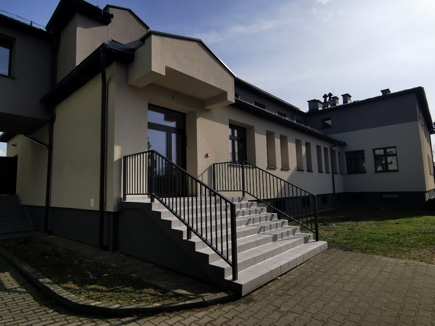 Widok zewnętrzny budynku z jasną elewacją. Na pierwszym planie schody z barierkami prowadzące do przeszklonych drzwi wejściowych. Po bokach ściany z rzędami prostokątnych okien.