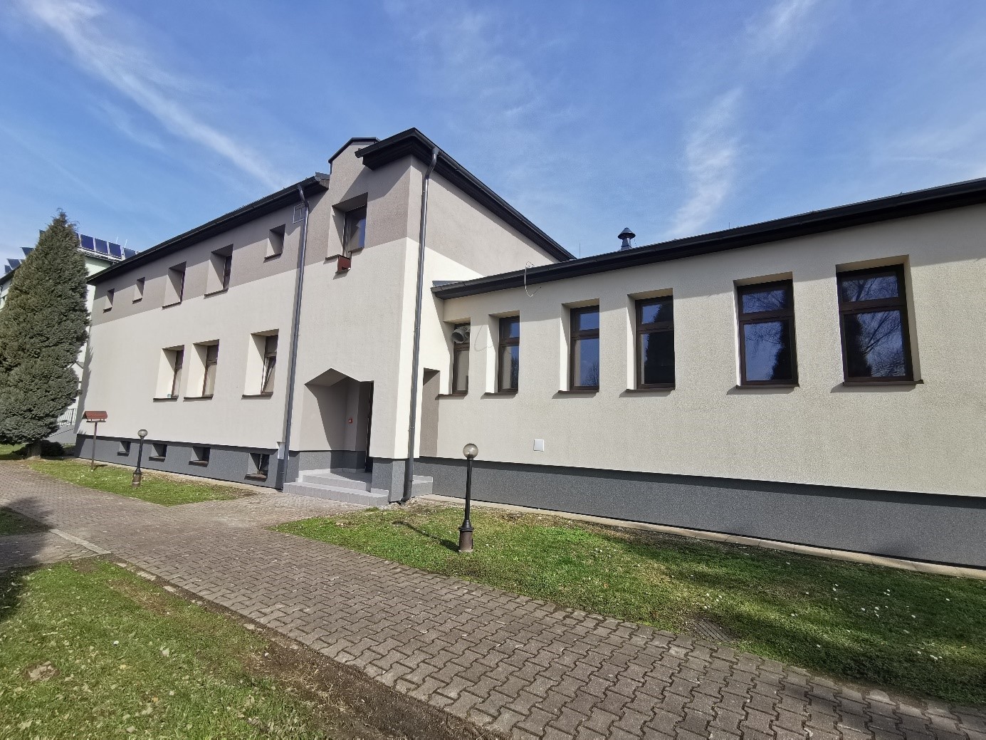 Budynek z jasną elewacją i ciemnymi ramami okiennymi, widoczny z boku. Na pierwszym planie chodnik z kostki brukowej i trawnik. Wejście prowadzi po kilku schodach bez barierki.