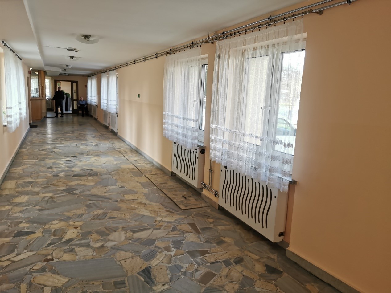 Wnętrze budynku z długim korytarzem, okna z firanami po prawej stronie, grzejniki pod oknami, podłoga z płytek o nieregularnych kształtach, na końcu korytarza dwie osoby.