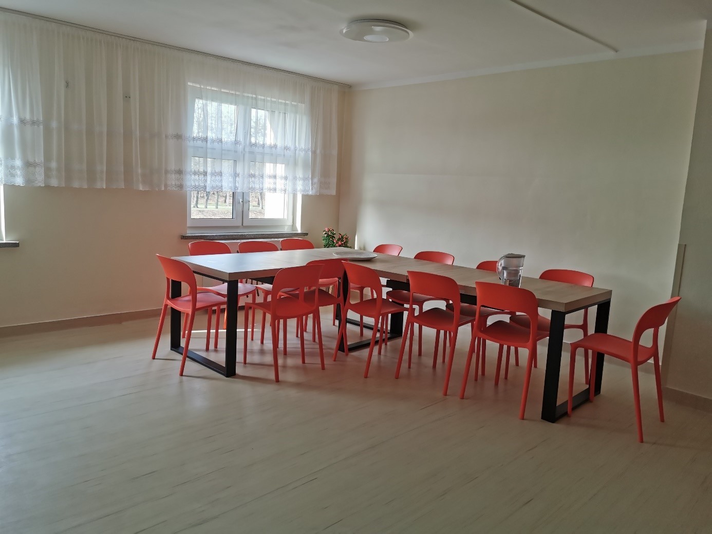 Jasne pomieszczenie z dużym oknem. W centralnej części dwa stoły ustawione w linii prostej, wok&oacute;ł nich czerwone krzesła. Na stole dzbanek z wodą. Podłoga jasna, ściany kremowe, okno zasłonięte firaną.