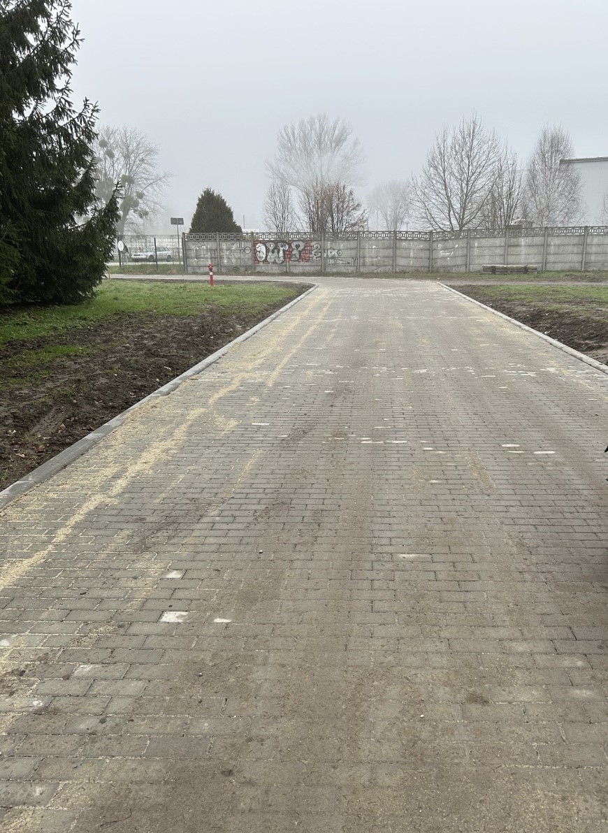 Nowo wybudowana droga z kostki brukowej prowadząca w stronę betonowego ogrodzenia z graffiti. Po bokach droga otoczona jest trawnikiem i ziemią. W tle widoczne drzewa i mglista pogoda.
