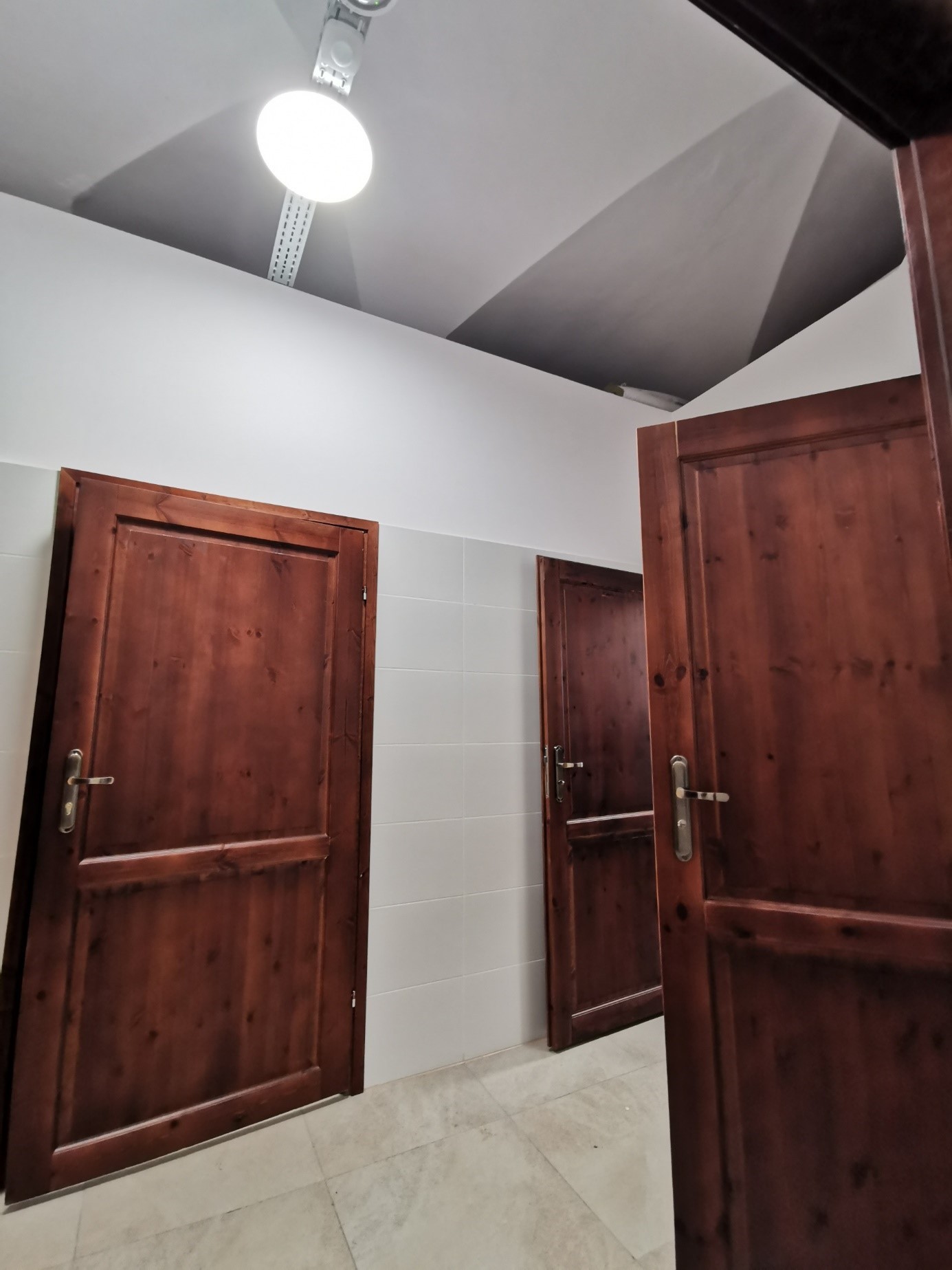 Wnętrze pomieszczenia sanitarnego z trzema drewnianymi drzwiami prowadzącymi do kabin. Ściany częściowo wyłożone białymi płytkami. Na suficie zamontowane oświetlenie sufitowe.