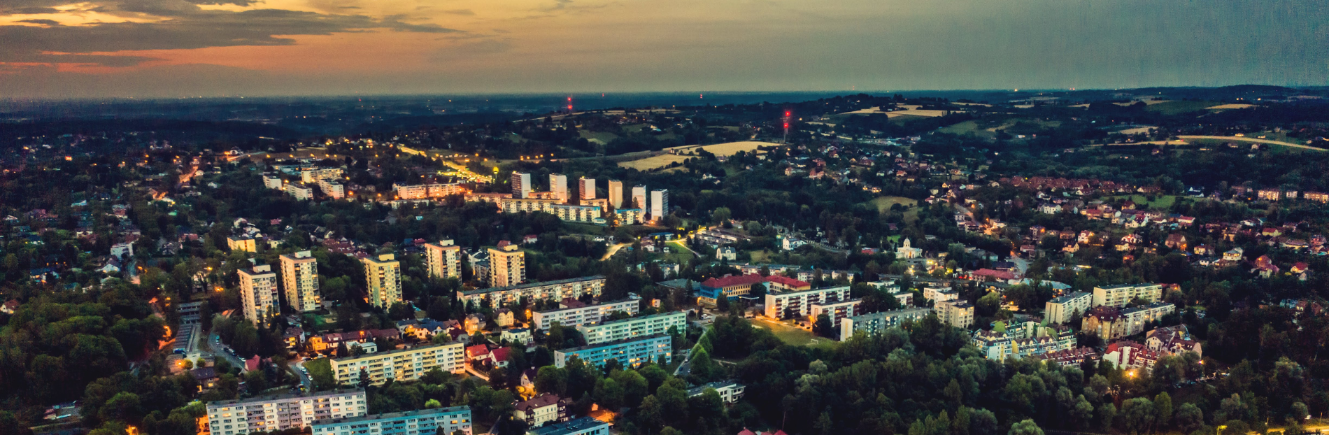 Panorama miasta o zmierzchu z widocznymi budynkami mieszkalnymi, ulicami i oświetleniem oraz otaczającymi terenami zielonymi.