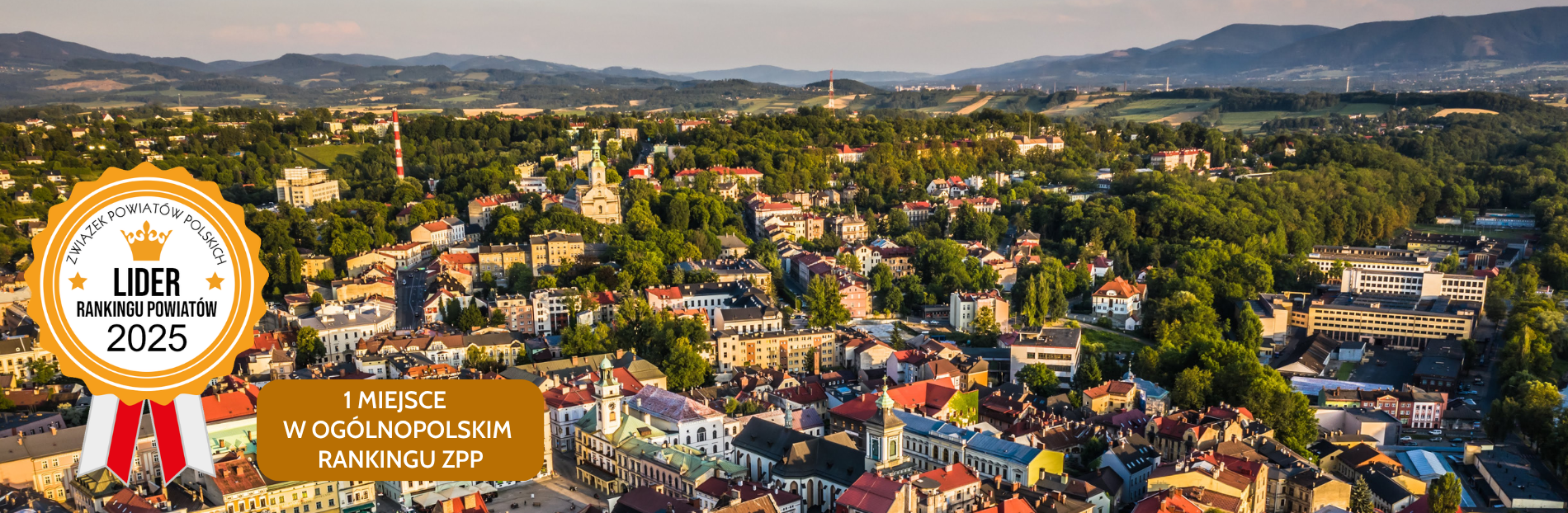 Widok miasta Cieszyn otoczonego zielenią i wzgórzami. W centrum historyczne budynki z czerwonymi dachami, wśród nich ratusz z wieżą. W tle góry i tereny wiejskie. Przestrzeń pełna drzew i parków, harmonijnie łącząca architekturę z naturą. 