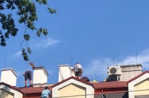 Widok na fragment dachu budynku podczas prac remontowych. Na dachu pracują osoby wykonujące prace budowlane, a poniżej ustawione jest rusztowanie. W tle widoczne są kominy oraz urządzenie techniczne zamontowane na dachu.