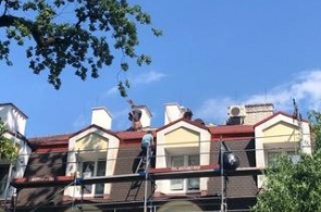 Widok na fragment budynku podczas prac remontowych na dachu. Przy elewacji ustawione jest rusztowanie, a na połaci dachowej pracują osoby wykonujące prace budowlane. W tle widoczne są kominy, okna dachowe oraz urządzenia techniczne.