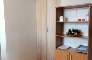 Pomieszczenie z jasną ścianą i drewnianą podłogą. Po lewej stronie szafa wnękowa z przesuwnymi drzwiami. Po prawej stronie regał z półkami i szafką dolną, na półkach dokumenty i dekoracje.