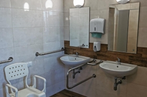 Pomieszczenie sanitarne z dwoma umywalkami i lustrami. Obok uchwyty wspomagające oraz plastikowe krzesło z oparciem i podłokietnikami. Na ścianie dozownik mydła i suszarka. Podłoga z płytek drewnopodobnych.