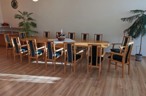 Pomieszczenie z dużym stołem i dwunastoma drewnianymi krzesłami z tapicerowanymi siedziskami. Podłoga wyłożona panelami. W tle dwie rośliny doniczkowe i dwie wiszące lampy.