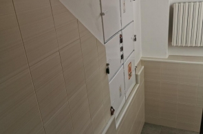 Klatka schodowa z szarymi stopniami i ścianą wyłożoną jasnymi płytkami. Na ścianie szafki instalacyjne, nad nimi okno i grzejnik. W górnej części ściany widoczne kolejne wnęki techniczne.