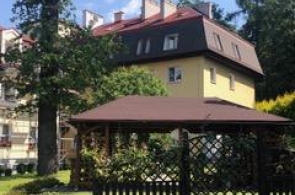 Widok na budynek mieszkalny otoczony zielenią. Na pierwszym planie znajduje się drewniana altana, a w tle widoczny jest budynek z czerwonym dachem, kominami i oknami dachowymi. Całość otaczają drzewa, trawnik i krzewy.