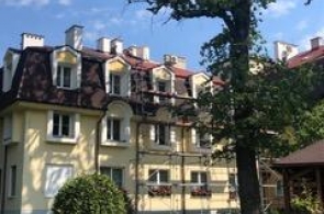 Widok na budynek mieszkalny otoczony zielenią. Na dachu widoczne są liczne kominy i lukarny, a przed budynkiem znajduje się trawnik z drzewami i krzewami.