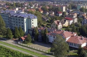 „Widok z lotu ptaka na kompleks budynków zlokalizowany w zabudowie miejskiej. Na pierwszym planie niższe budynki z czerwonymi dachami, w tle wysoki budynek z panelami fotowoltaicznymi na dachu.