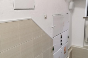 Klatka schodowa z szafkami instalacyjnymi zamontowanymi na ścianie. Ściana częściowo wyłożona jasnymi płytkami. Na podłodze widoczne schody z zielonym stopniem.
