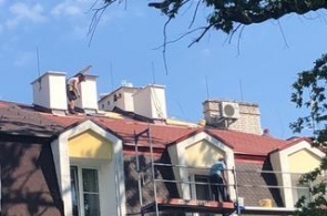 Widok na fragment budynku podczas prac remontowych dachu. Na połaci dachowej pracują osoby wykonujące prace budowlane, a przy elewacji ustawione jest rusztowanie. W tle widoczne są kominy, lukarny oraz urządzenie techniczne zamontowane na dachu.