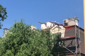 Widok na fragment budynku podczas prac remontowych. Przy elewacji ustawione jest rusztowanie, a na dachu widoczne są elementy konstrukcyjne i urządzenia techniczne. Dolną część budynku częściowo zasłaniają drzewa.