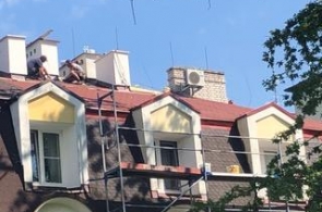 Widok na fragment budynku podczas prac remontowych dachu. Na połaci dachowej pracują osoby wykonujące prace budowlane, a przy elewacji ustawione jest rusztowanie. W tle widoczne są kominy, lukarny oraz urządzenie techniczne zamontowane na dachu.