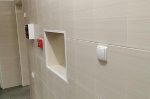 Fragment korytarza z jasnymi płytkami na ścianie i szarą podłogą. Na ścianie wnęka, przycisk alarmowy, czujnik i skrzynka instalacyjna. U góry widoczny fragment instalacji sufitowej.