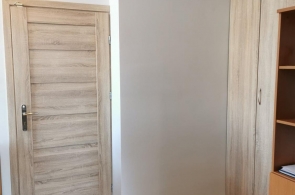 Fragment wnętrza pomieszczenia z jasnymi ścianami, drewnianymi drzwiami, szafą wnękową z przesuwnymi drzwiami i częścią regału. Podłoga wyłożona panelami.