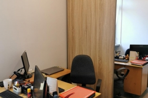 Pomieszczenie biurowe z dwoma stanowiskami pracy. Na biurkach komputery, dokumenty i przybory biurowe. Pośrodku przesuwane drzwi z jasnego drewna. Na podłodze panele, w tle okno.