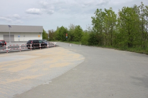 Widok na utwardzony plac przy drodze, częściowo pokryty piaskiem. Po lewej stronie znajduje się parking z samochodami oraz barierki, a w tle widoczne są znaki drogowe, budynek i otaczająca teren zieleń.