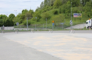 Widok na rozległy, utwardzony plac przy drodze, częściowo pokryty piaskiem. Wzdłuż krawędzi ustawiono metalowe stojaki oraz znaki drogowe. W tle widoczna jest jezdnia, samochód ciężarowy i porośnięte zielenią zbocze.