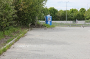 Widok na utwardzony plac otoczony zielenią. Po lewej stronie rosną drzewa i krzewy, a w tle znajduje się przenośna toaleta, ogrodzenie oraz elementy infrastruktury drogowej.