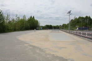 Widok na utwardzony plac przy drodze, częściowo pokryty piaskiem. Teren jest oddzielony barierkami, a obok znajdują się latarnie z panelami solarnymi. W tle widoczna jest jezdnia, zieleń oraz pojedyncze pojazdy.
