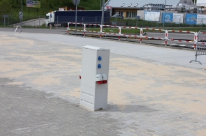 Na utwardzonym placu znajduje się skrzynka z gniazdami przyłączeniowymi. W tle widoczne są barierki oddzielające teren od jezdni, samochód ciężarowy oraz zabudowania usługowe. Część nawierzchni placu pokryta jest piaskiem.