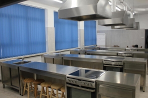 Pracownia gastronomiczna z nowoczesnym wyposażeniem; stalowe blaty robocze, kuchenki, wysokie okapy oraz niebieskie rolety w oknach, sala w ZSGH w Wiśle.