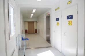 Stanowisko komputerowe w pracowni diagnostycznej z dwoma monitorami i sprzętem sterującym, za szybą widoczny tomograf komputerowy.