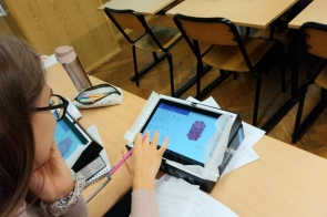 Uczeń lub uczennica siedzi przy ławce i obsługuje tablet z rysunkiem bryły 3D na ekranie, obok leżą przybory szkolne i kartki.