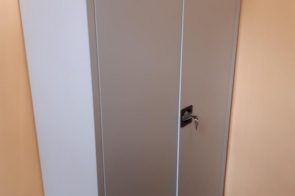 Zamknięta metalowa szafa z dwoma znakami ostrzegawczymi o substancjach niebezpiecznych, ustawiona przy ścianie w pomieszczeniu.
