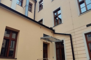 Tylna część budynku szkoły II liceum ogólnokształcącego w Cieszynie z wejściem po schodach, poręczą i zadaszeniem. Widoczne wysokie okna i jasna elewacja.