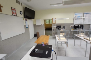 Sala lekcyjna z białą tablicą, projektorem, biurkiem nauczyciela i metalowymi stanowiskami uczniowskimi; na ścianie krzyż, zegar i tablice informacyjne - sala w ZSTiO w Skoczowie.