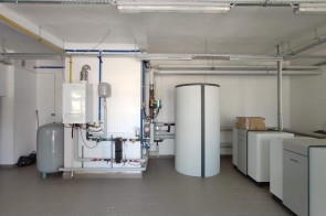 Pomieszczenie techniczne z instalacją grzewczą, zbiornikami i urządzeniami kotłowni. Widoczne rury, zbiornik buforowy i moduły grzewcze w pomieszczeniu Centrum Kształcenia Zawodowego w Bażanowicach.