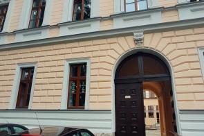 Fasada zabytkowego budynku szkoły II liceum ogólnokształcącego w Cieszynie z ozdobnymi oknami i łukowym wejściem. Przed wejściem zaparkowane samochody na brukowanym podjeździe.