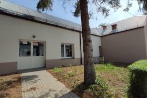 Budynek o jasnej elewacji z wejściem przez przeszklone drzwi. Przed wejściem chodnik i trawnik z drzewem oraz żywopłotem przy Centrum  Kształcenia  Zawodowego w Bażanowicach.