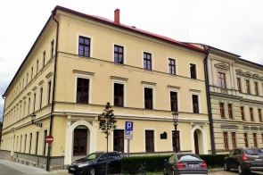 Zabytkowy budynek szkoły Zespołu Szkół Budowlanych  w Cieszynie z jasną elewacją i czerwonym dachem. Trzypiętrowa kamienica narożna z zaparkowanymi samochodami przy ulicy.
