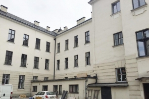 Widok wewnętrznego dziedzińca budynku szkolnego I liceum ogólnokształcącego w Cieszynie z jasną elewacją i wieloma oknami. Na dole zaparkowane pojazdy i drzwi techniczne.