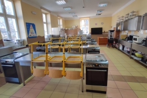 Sala dydaktyczna z wyposażeniem kuchennym, stanowiskami z piekarnikami, zlewami i żółtymi krzesłami. Po prawej stronie ciąg blatów z mikrofalówkami i szafkami wiszącymi w Zespole Placówek Szkolno-Wychowawczo-Rewalidacyjnych w Cieszynie.