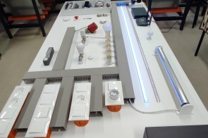 Stół z układem do nauki instalacji elektrycznej, zawierającym moduły, włączniki, żarówkę i oprawy świetlówkowe. W tle stanowiska dydaktyczne w Zespole Szkół Technicznych w Cieszynie.